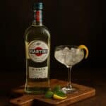 Martini Bianco in bottiglia su tagliere di legno con bicchiere ghiacciato, scorza di limone e foglia di menta