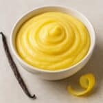 Crema pasticcera liscia in ciotola con baccello di vaniglia e scorza di limone su sfondo chiaro