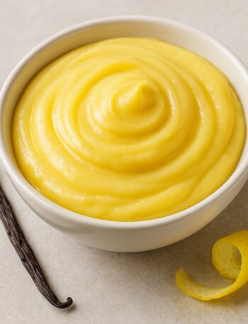 Crema pasticcera liscia in ciotola con baccello di vaniglia e scorza di limone su sfondo chiaro