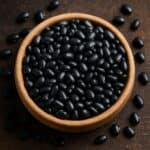 Fagioli neri lucidi in ciotola di legno chiaro su tavolo scuro con alcuni chicchi sparsi