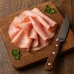 Fette di mortadella rosa su tagliere in legno con coltello e pistacchi verdi accanto