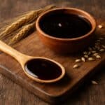 Malto liquido di colore ambrato scuro in una ciotola di legno e cucchiaio, con chicchi d’orzo e spighe su tagliere