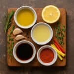 Vari ingredienti per marinature disposti su un tagliere rustico con olio, erbe aromatiche, spezie, limone e aglio
