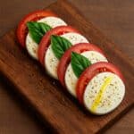Insalata Caprese: Mozzarella, pomodoro e basilico disposti in sequenza su tagliere con olio e pepe