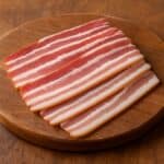 Fette strette di pancetta con venature regolari su tagliere di legno rotondo