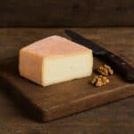 Fetta di taleggio a forma rettangolare con crosta rosata su tagliere in legno, accanto a noci e coltello