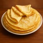 Crepes dolci e salate impilate su piatto bianco, con una crêpe piegata in primo piano su sfondo caldo in legno.