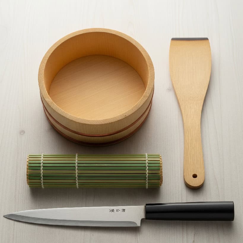 Visti dall'alto, gli strumenti principali per preparare il sushi: una ciotola di legno (hangiri), una spatola di legno (shamoji), una stuoia di bambù (makisu) e un coltello da sushi affilato.