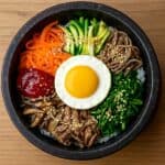 Bibimbap servito in una ciotola di pietra nera con riso, verdure colorate, carne, uovo al tegamino e gochujang, visto dall’alto.
