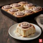 Un cinnamon roll singolo e perfettamente glassato su un piatto bianco, con una teglia di altri rolls appena sfornati sfocata sullo sfondo.