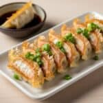 Gyoza dalla base dorata e croccante disposti su un piatto bianco rettangolare, con una sottile ala di farina che li unisce. Accanto, una ciotola con la salsa d'accompagnamento.