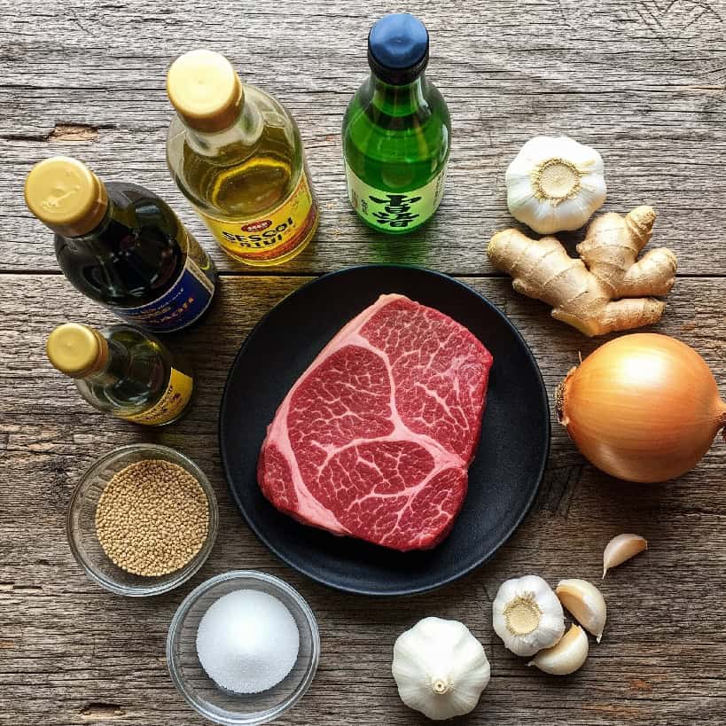 Disposizione dall'alto degli ingredienti per Bulgogi: una fetta di manzo crudo, cipolla, aglio, zenzero, salsa di soia, olio di sesamo e zucchero su un tavolo di legno rustico.