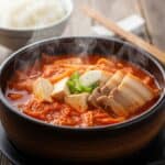 Una ciotola fumante di Kimchi Jjigae con stufato rosso, cubetti di tofu, fette di maiale e kimchi, servita su un tavolo di legno scuro.