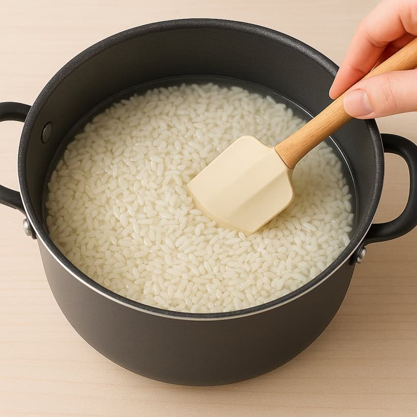 Riso per sushi versato in una pentola antiaderente con acqua, livellato con una spatola per garantire una cottura uniforme.