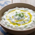 Una ciotola di legno piena di Tzatziki cremoso con cetriolo e aneto, su un tavolo di legno.