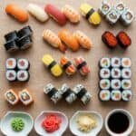 Visti dall'alto, diversi tipi di sushi fatti in casa (nigiri, maki, uramaki) disposti su un tavolo di legno chiaro, con wasabi, zenzero marinato e salsa di soia.