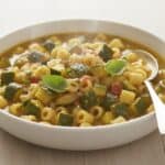 Fotografia di una ciotola di pasta e zucchine, con pasta corta e cubetti di zucchina. Il piatto è guarnito con foglie di basilico e un cucchiaio.