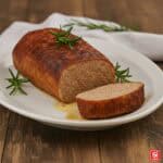 Fotografia di un polpettone al forno su un tagliere in legno scuro. Una fetta è stata tagliata e spostata leggermente, rivelando la sua consistenza interna. La superficie del polpettone ha una crosta dorata e lucida.