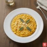 Immagine dall'alto di un risotto alla zucca cremoso in un piatto piano bianco, guarnito con Parmigiano grattugiato e salvia fresca.