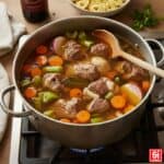 Pentola di brodo di carne fumante con pezzi di manzo, carote, sedano, cipolla rossa e spezie, cotto su un fornello a gas. Si intravede una bottiglia di vino rosso e una ciotola di tortellini sullo sfondo.