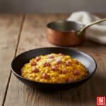 Risotto alla Monzese cremoso all'onda con Luganega di Monza, zafferano e Grana Padano, in ciotola di ceramica scura.