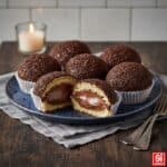 Pasticcini Teste di Moro Napoletane, ricoperti da codette di cioccolato e serviti in pirottini bianchi. Una monoporzione è tagliata a metà, mostrando il ripieno di morbida crema al cioccolato.