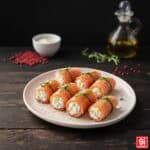 Involtini di salmone affumicato ripieni di crema di ricotta e limone, legati con erba cipollina e decorati con pepe rosa su un piatto bianco moderno.