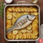 Orata al forno con patate croccanti servita in teglia, guarnita con rosmarino e aromi.