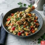 Cous cous con pollo e verdure colorate, servito in bowl su marmo chiaro, con forchetta che solleva un boccone.