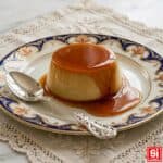 Crème caramel intero con caramello ambrato su piatto elegante decorato blu e oro con cucchiaio d’argento su piano chiaro.